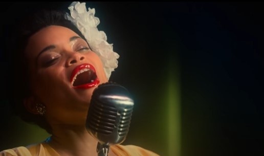 Andra Day as Billie Holiday, leaning her head back to sing into a microphone. Her mouth is open and she wears a large white flower on one side of her head, and glossy red lipstick on her lips