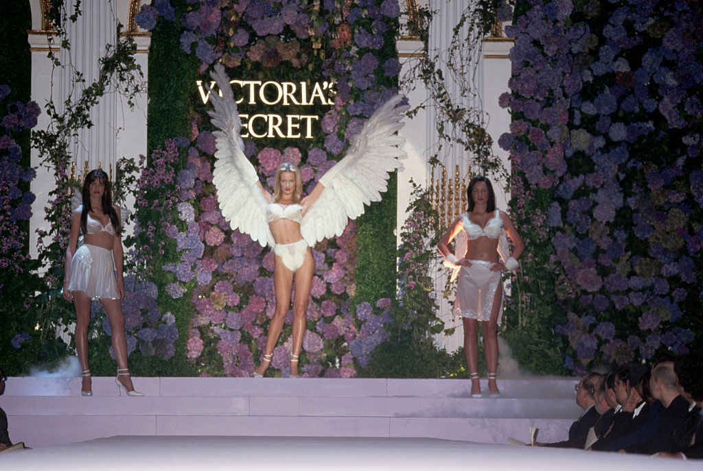 Three models at a Victoria&#x27;s Secret fashion show, the middle of which is wearing giant wings on her arms