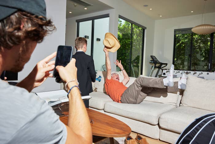 An older man clutching a panama hat falls backward onto a couch while a younger person in the foreground records it on a phone