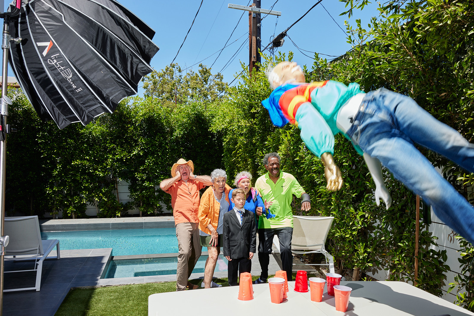 Older people and a young boy in an 8-year-old stand outside and react as a body doll falls onto a table with red solo cups