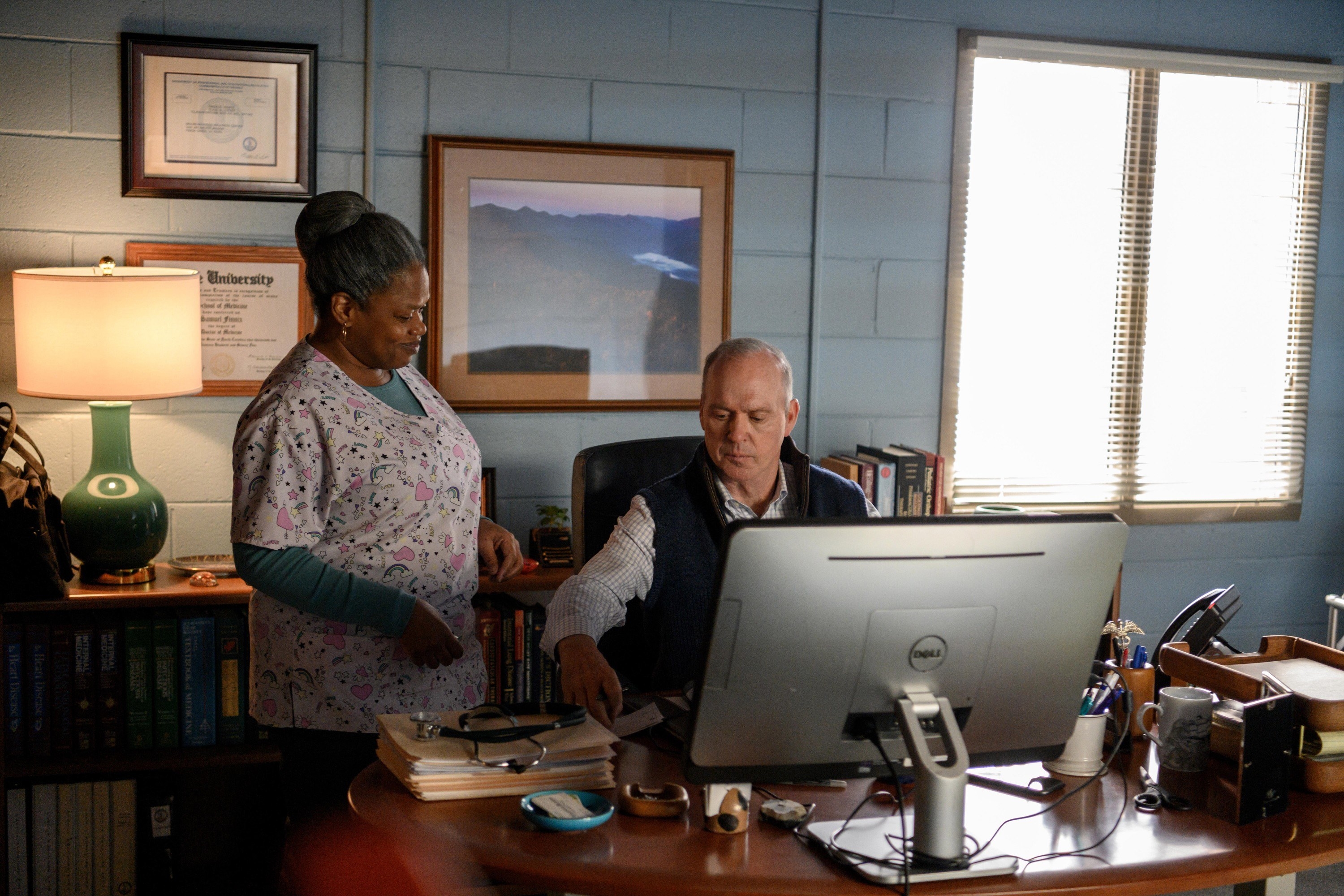 Michael Keaton as Dr Samuel Finnix sitting at a desk with a computer on it and Arischa Conner as Leah Turner standing next to him