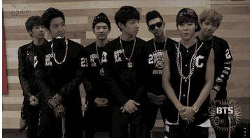A gif from 2013: The 7 members of BTS stand in two rows wearing black and white clothes and large gold chains