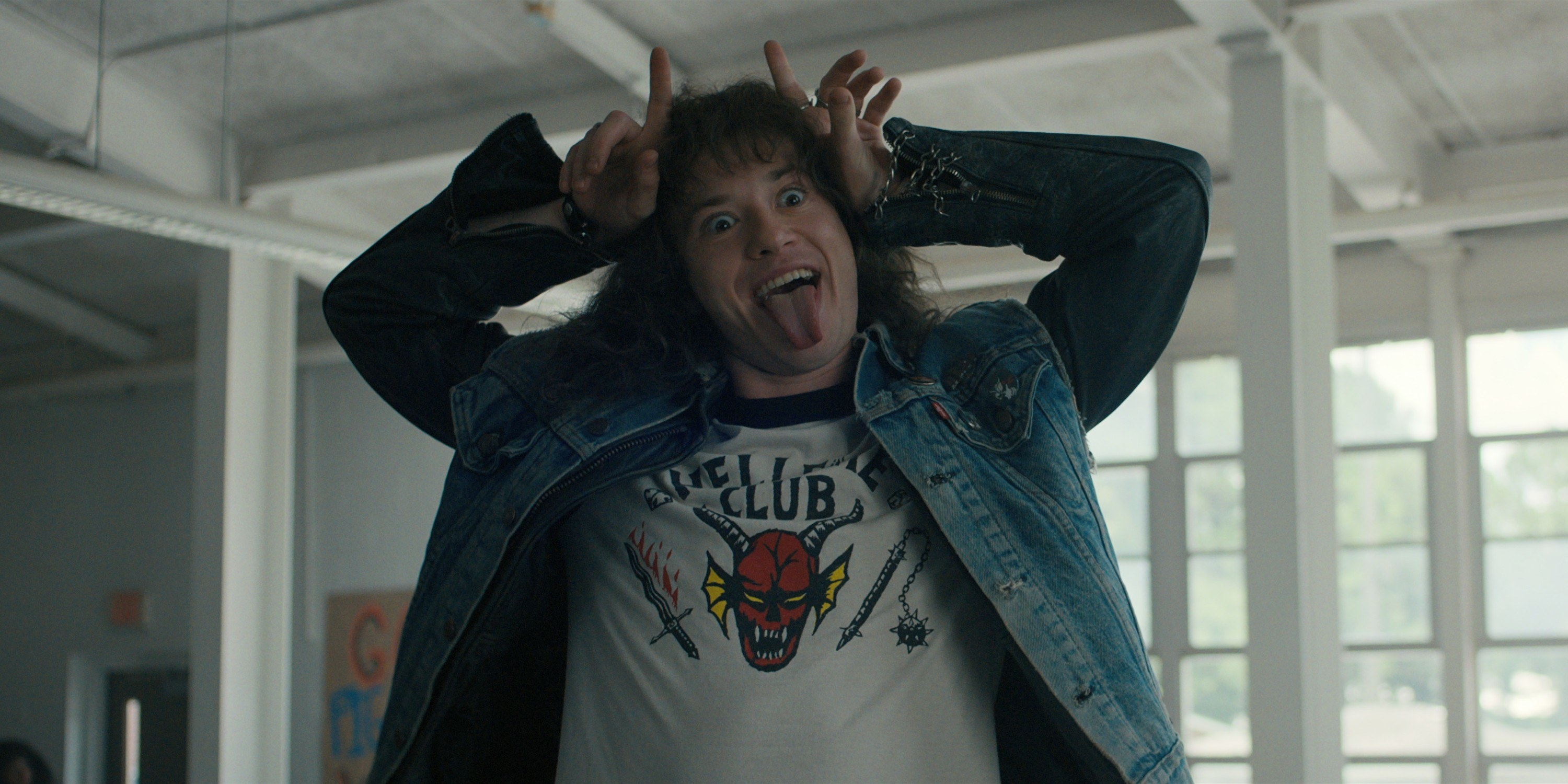 Eddie Munson wears a Hellfire Club T-shirt under a denim vest while making devil horns with his fingers and his tongue sticking out