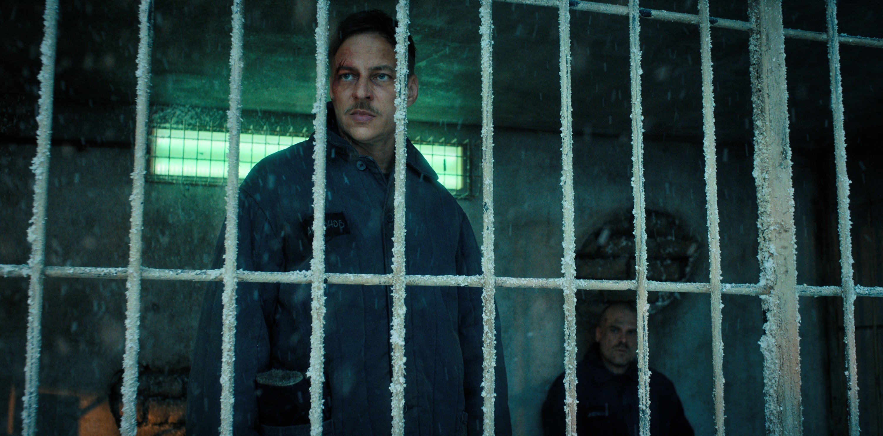 Dmitri wears a dark jumpsuit as he stands behind prison bars