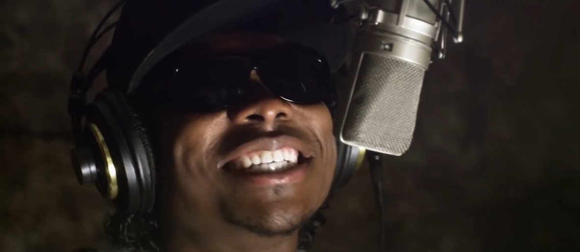 A rapper in the recording studio, wearing headphones, a baseball cap and sunglasses. The microphone is in front of his face and he is mid-rap, his mouth open