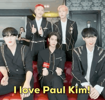 Half of CIX are standing up and half are sitting on a sofa while Seunghun 승훈 is making a love heart with his hands saying he loves singer Paul Kim