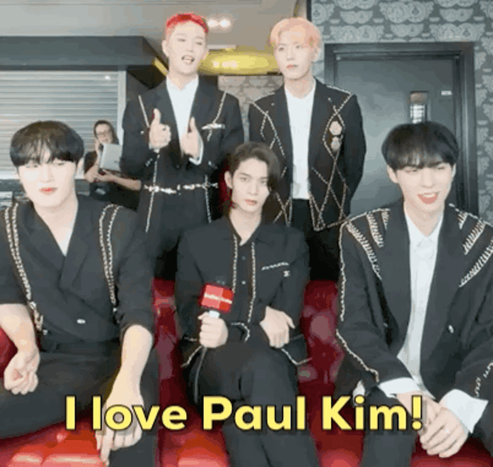 Half of CIX are standing up and half are sitting on a sofa while Seunghun 승훈 is making a love heart with his hands saying he loves singer Paul Kim
