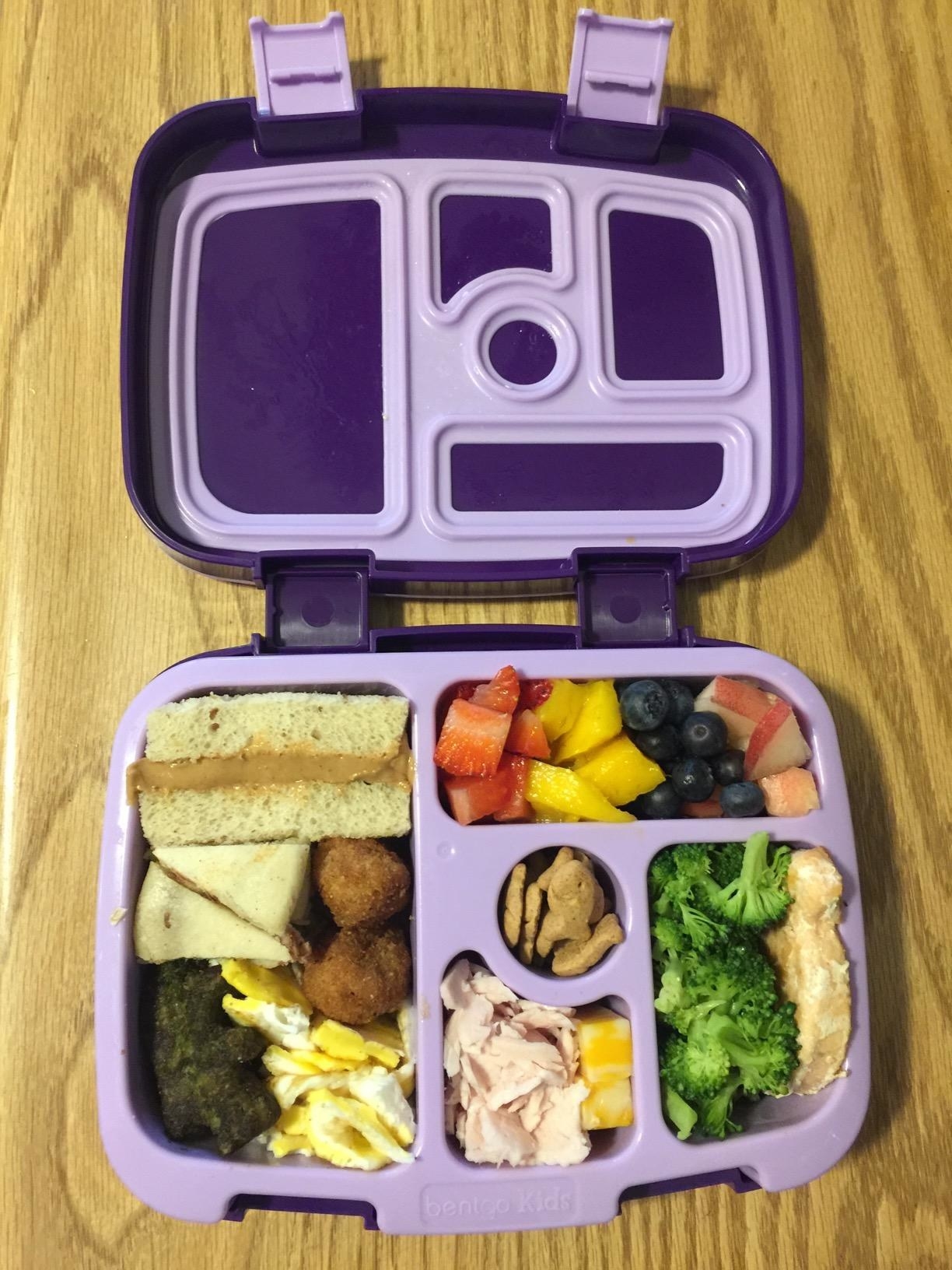reviewer&#x27;s photo showing the purple bento box open and packed for their kid with peanut butter sandwich, egg, turkey and cheese, broccoli and chicken, fruit, and graham crackers among the various compartments