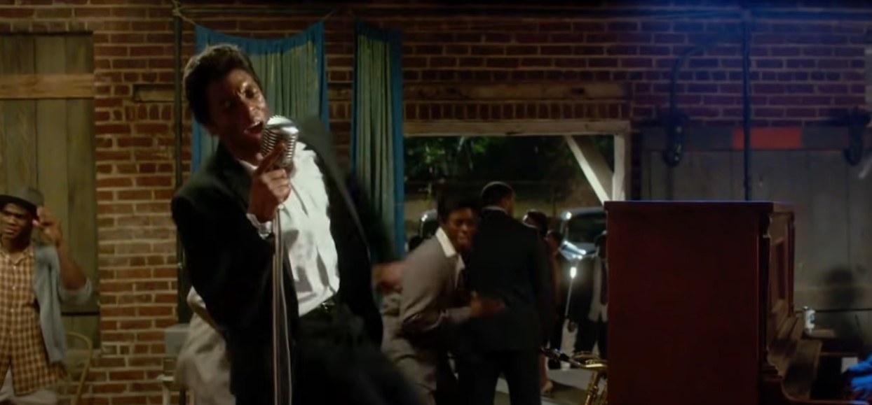 Chadwick Boseman as James Brown, wearing a black and white tux and leaning back gripping an old fashioned microphone as he sings expressively