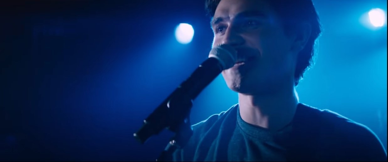 KJ Apa as Jeremy Camp, singing into a mic on stage with blue lights illuminating him from behind