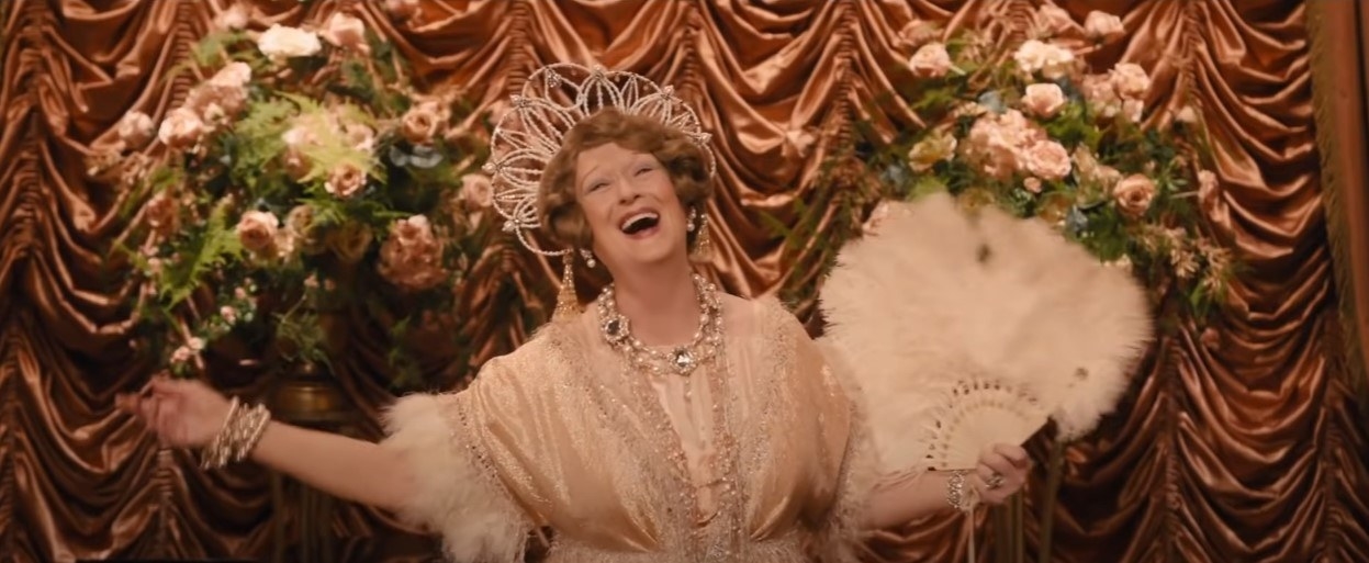 Meryl Streep as Florence Foster Jenkins, singing with her arms outstretched with a white feather fan in one, in front of a rust coloured draped velvet backdrop