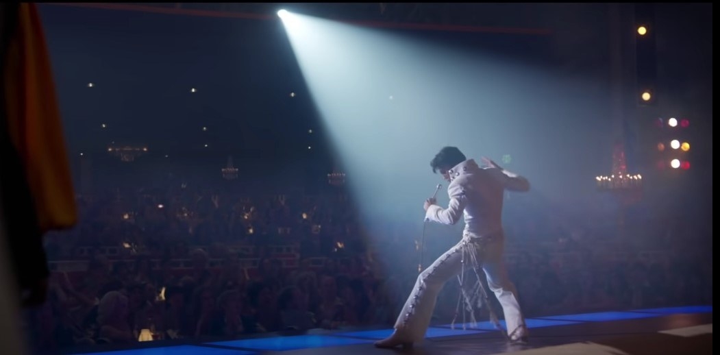 Austin Butler as Elvis, wearing a white leather catsuit and performing onstage. The shot is taken from behind, showing a full audience in front of the performer