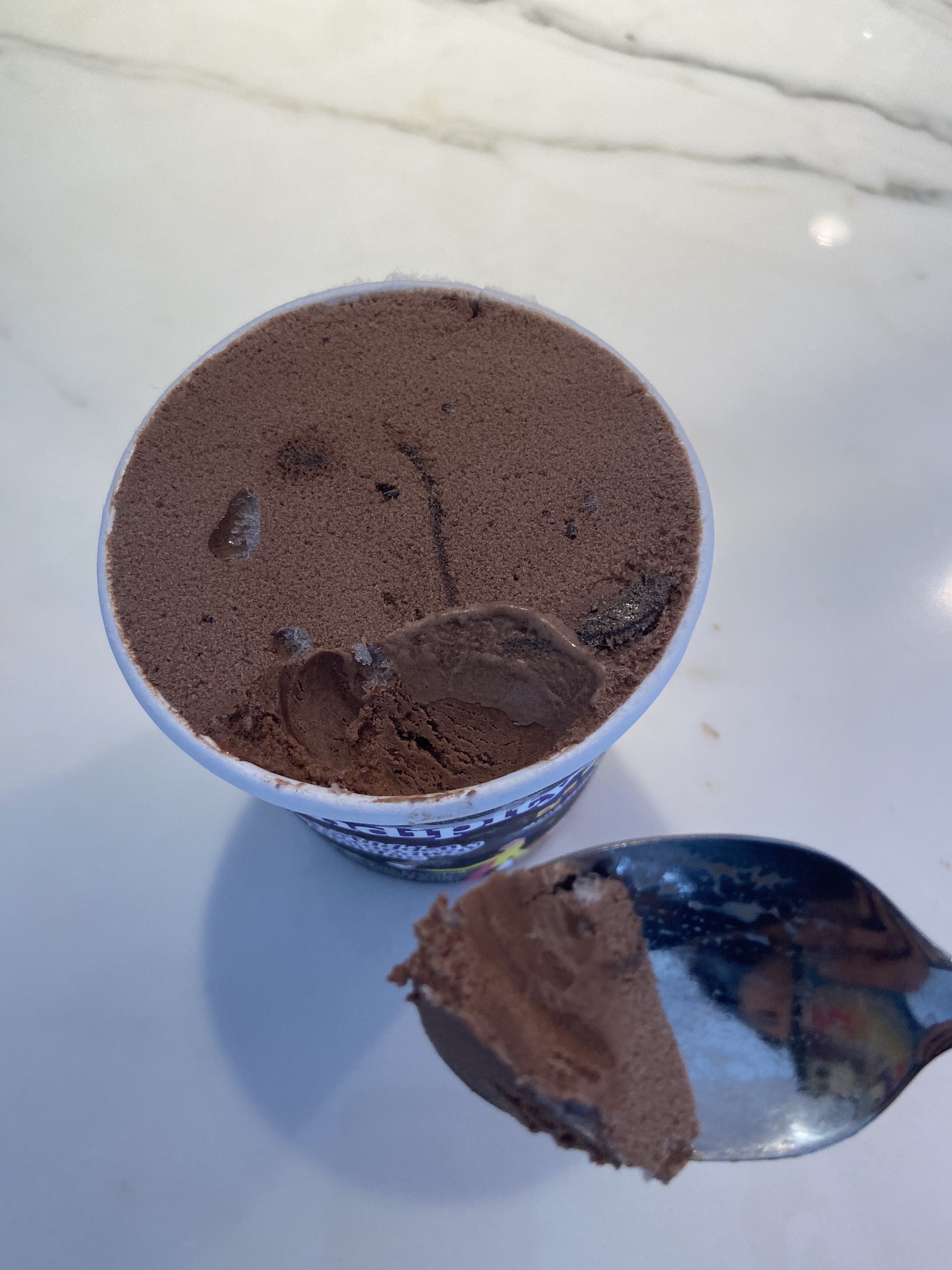 The ice cream opened and with one spoonful taken out; it is the color of chocolate, with a few visible chunks of brownie and one visible fudge swirl