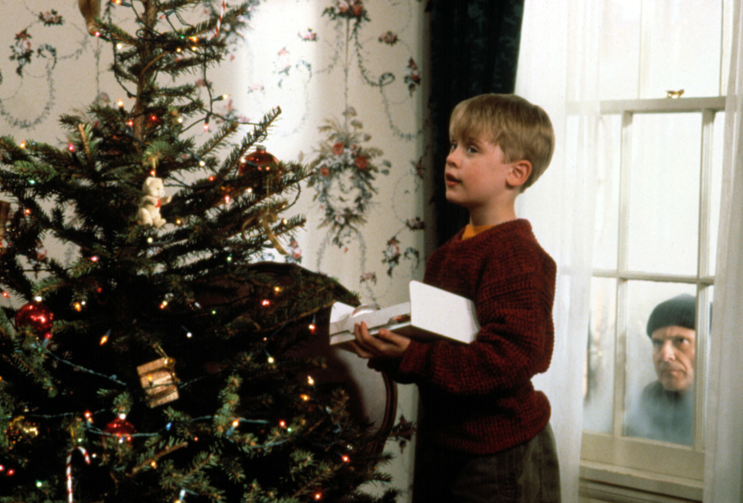 a kid stands by a Christmas tree as a man looks in the window