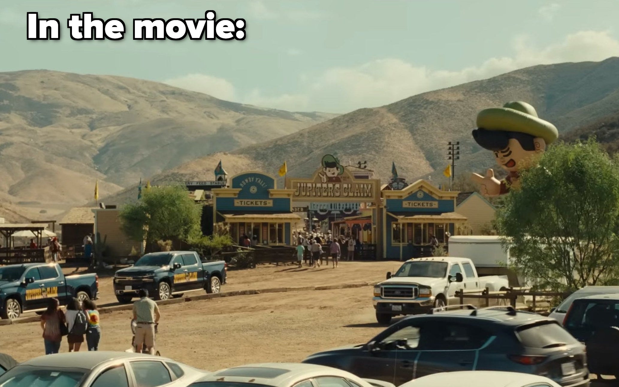 A photo of Jupiter&#x27;s Claim in the movie, with a large sign marking the entrance to the town, which has a giant inflatable mascot next to it and rolling hills behind it