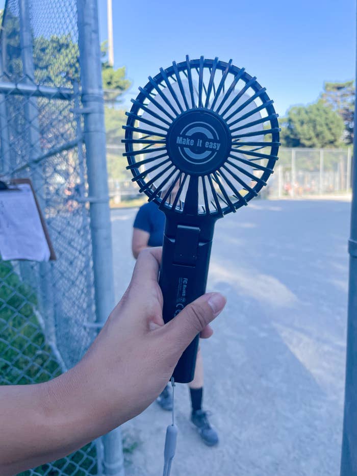 a person holding up the adjustable fan while at a baseball diamond