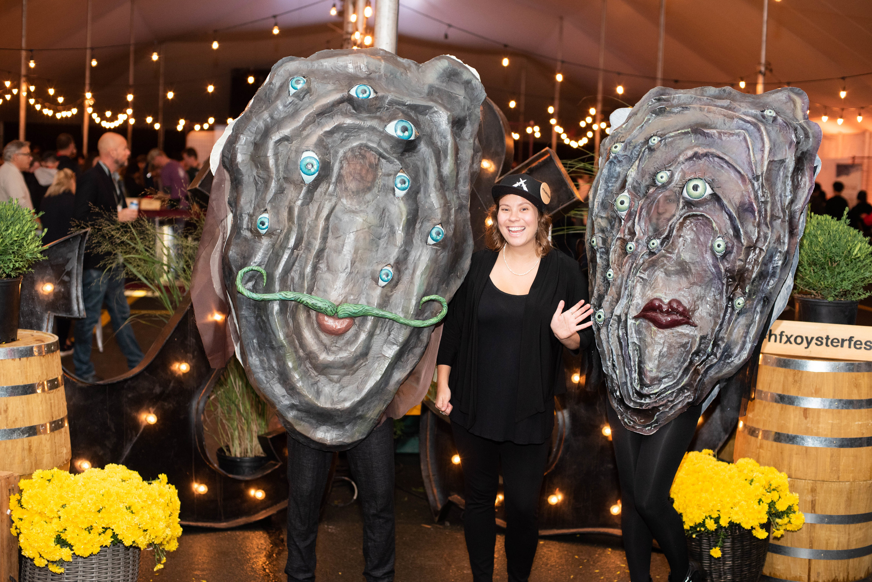 A woman waves hello standing between two oversize oyster costumes with a terrifying number of eyes and vacant expressions