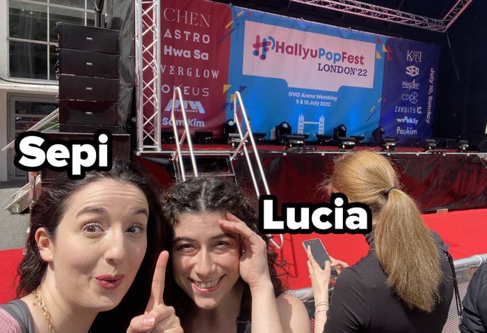 Sepi and Lucia from BuzzFeed UK stand in front of a stage which says HallyuPopFest London 22; Sepi is pointing up to the stage, and both look super excited