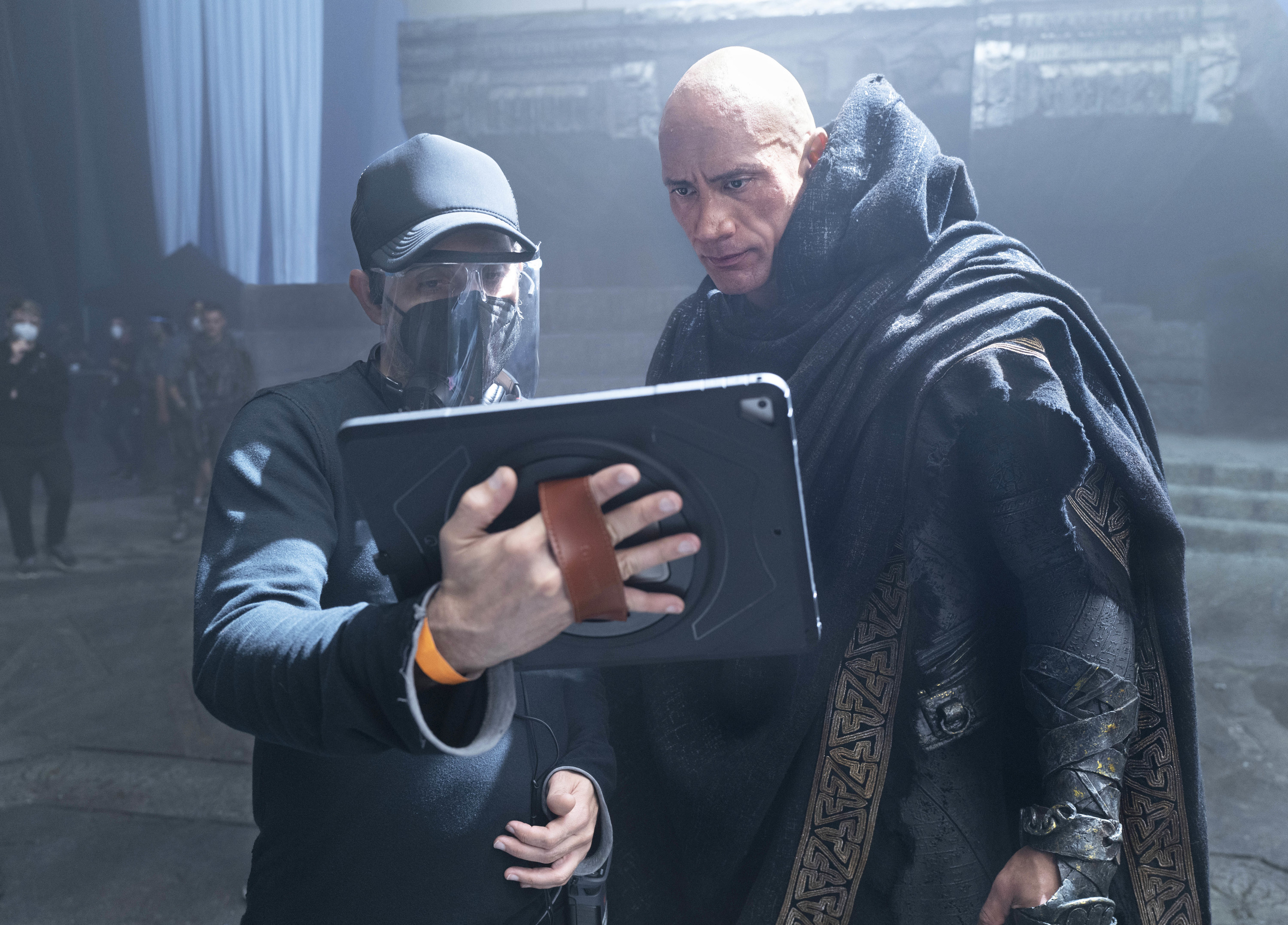 Jaume, wearing a face mask and additional face covering, shows something to Dwayne on a tablet on the set of Black Adam