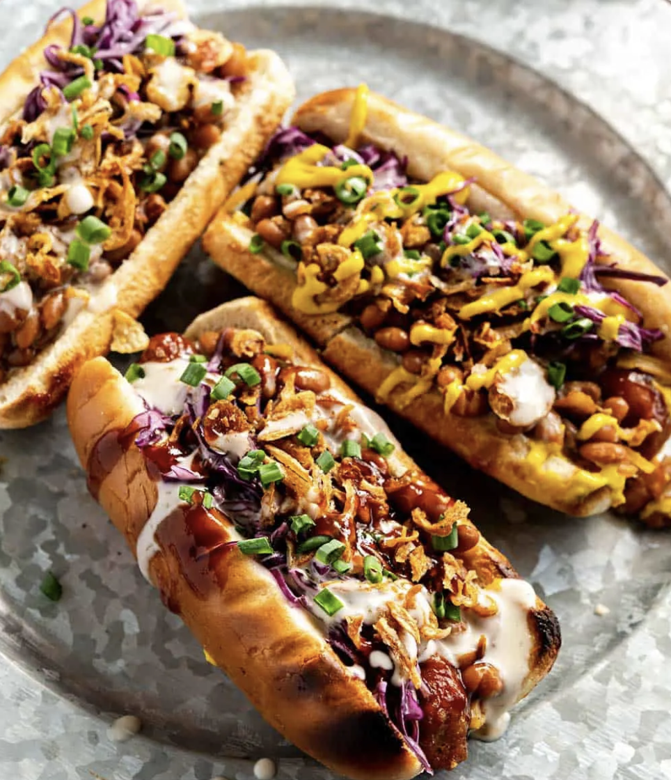 three vegan hot dogs with onions, scallions, cabbage, baked beans, and sauce all over the top