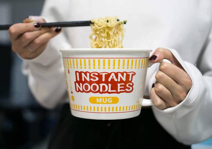 a person eating ramen noodles out of a big mug that says instant noodles mug on it