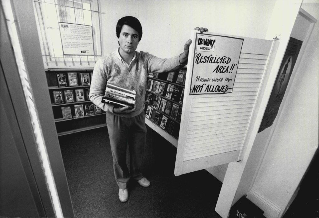 A man carries a stack of movies while leaning on a saloon door that says &quot;restricted area, persons under 18 years not allowed&quot;