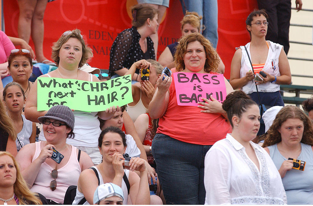 People in an audience with some holding signs, including &quot;Bass is back!&quot; and &quot;What&#x27;s next for Chris&#x27;s hair?&quot;