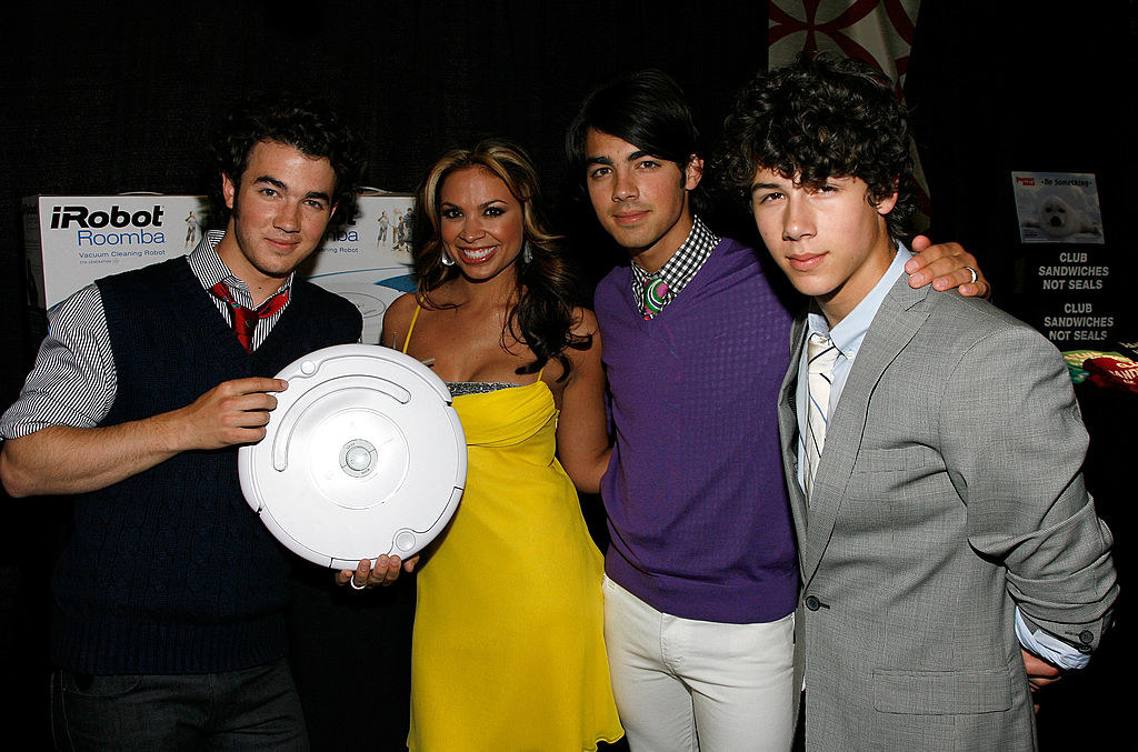 The Jonas Brothers standing next to a woman as one of them holds a Roomba