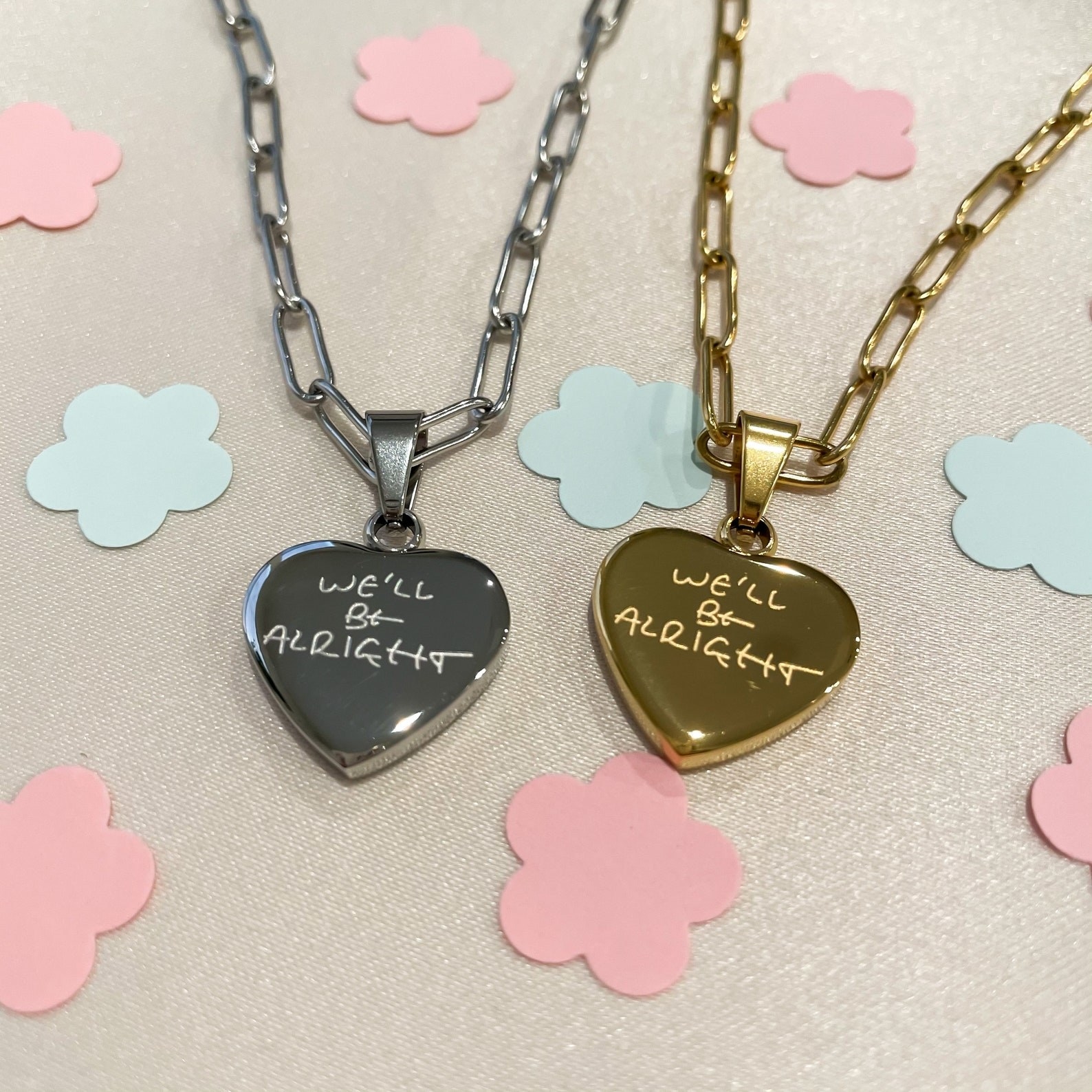 Silver and gold heart pendant side by side with engraving &quot;We&#x27;ll be alright&quot; on them