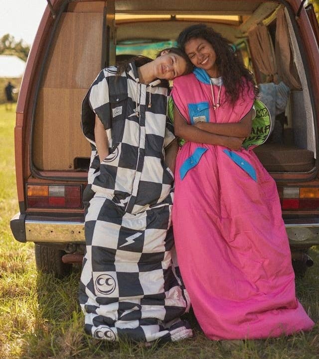 two people leaning against the back of a camper van wearing the sleeping bags