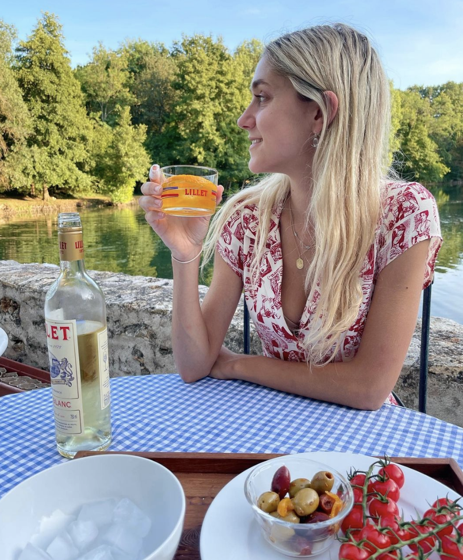 Claire sipping on Lillet in front of a body of water with olives and cherry tomatoes as a snack