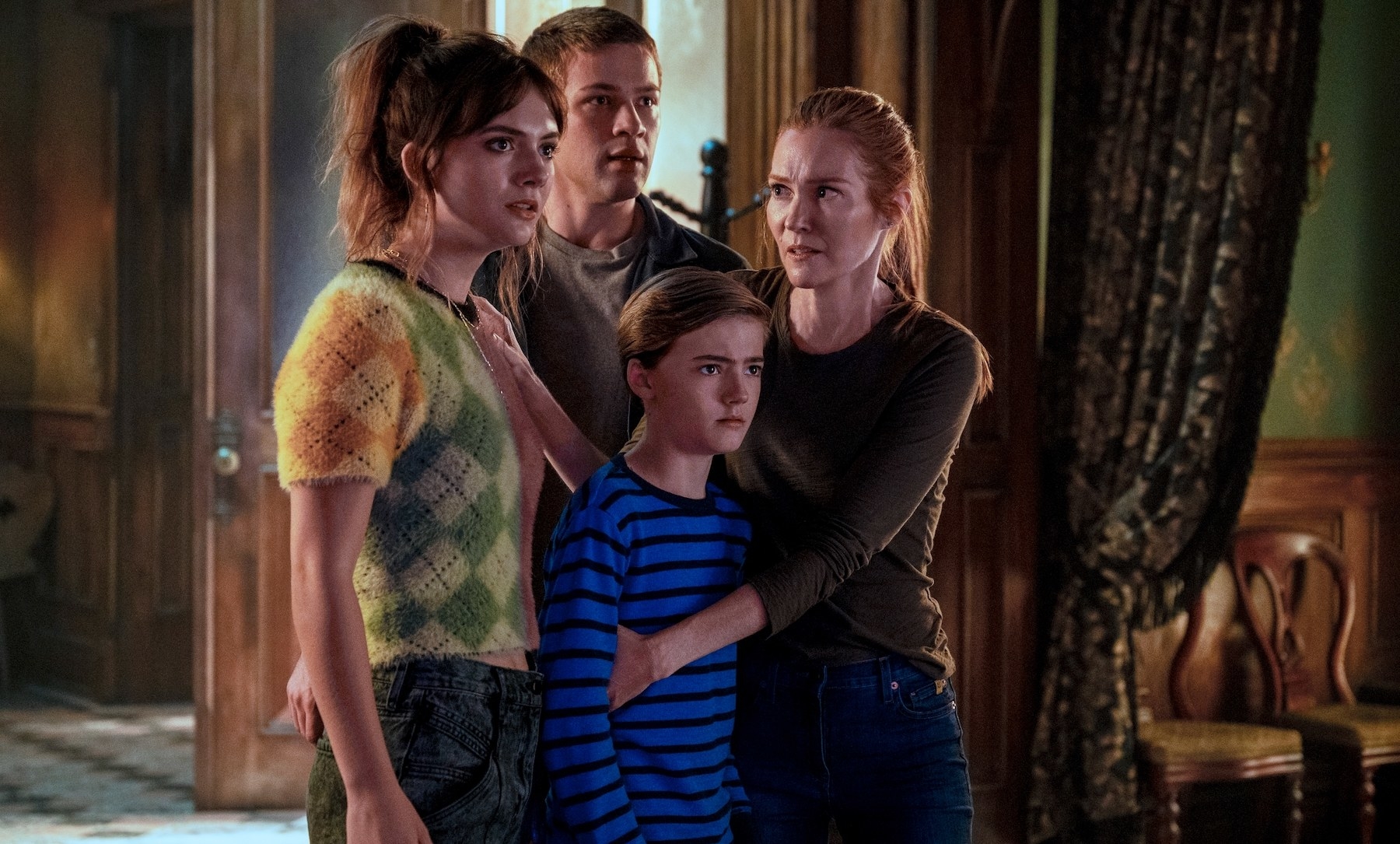 three kids and their mother huddle together looking concerned at something out of frame in the middle of a grand looking house