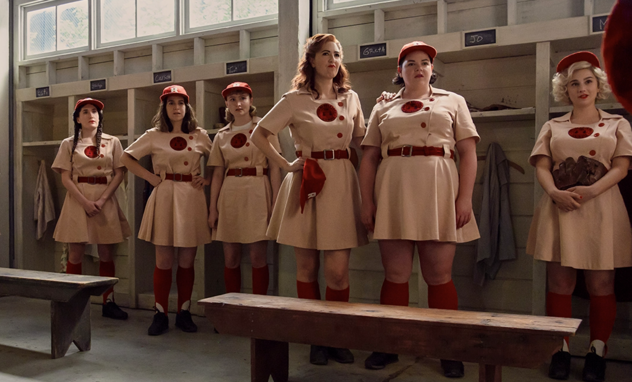 Several women in old fashioned baseball uniforms group together in a locker room