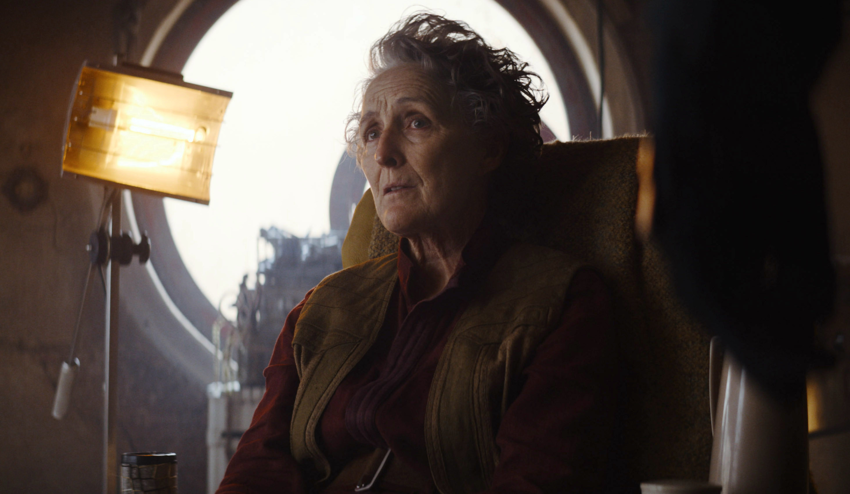 An old woman played by fiona shaw sits in an office with a lamp and a round window and looks at something out of frame with a disconcerted look