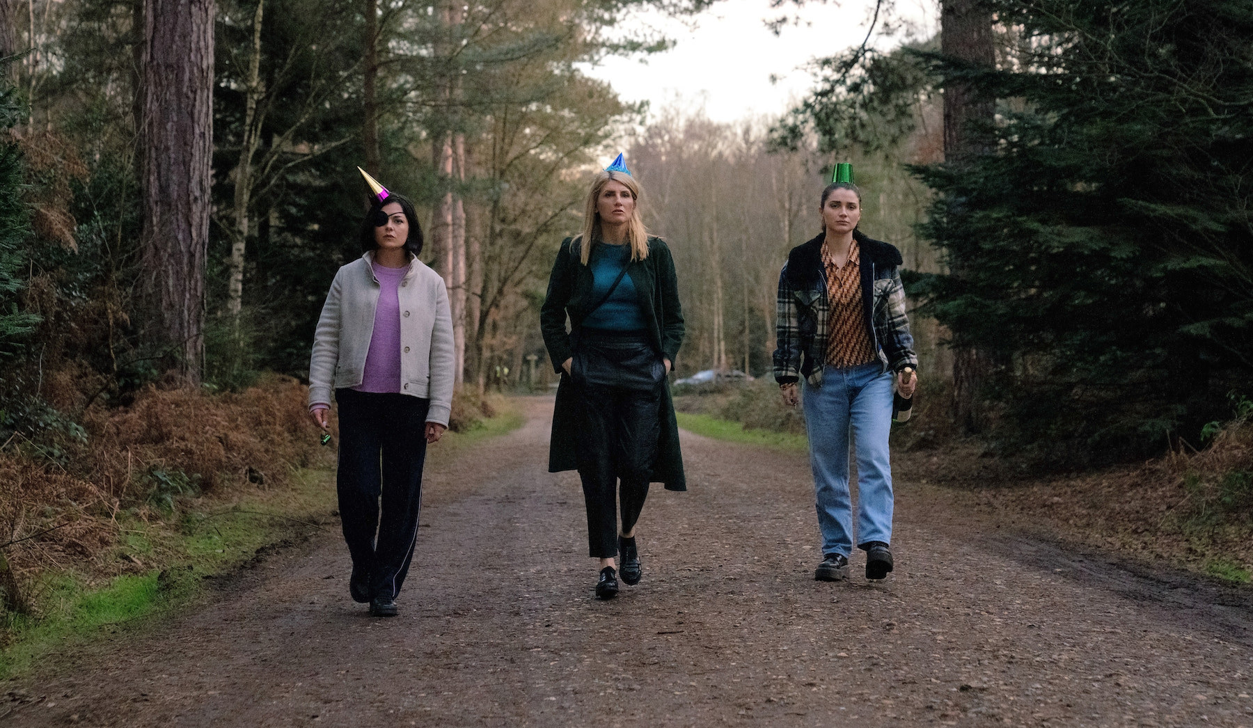 Three adult women walk down an empty road in forest side by side wearing party hats