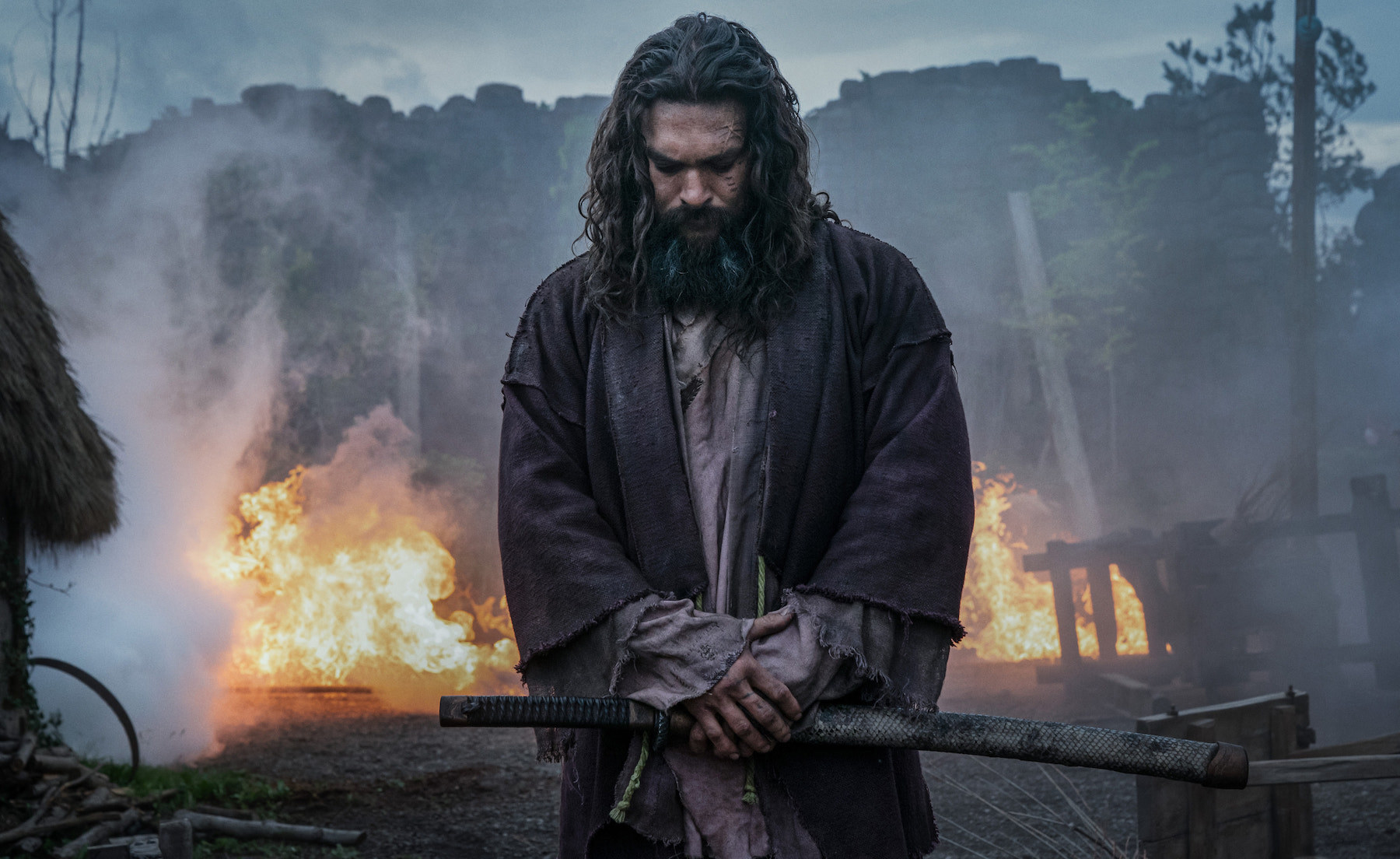 A man played by jason momoa in tattered robes stands in a burning forest with a samurai sword in his hands and looks down as if in prayer