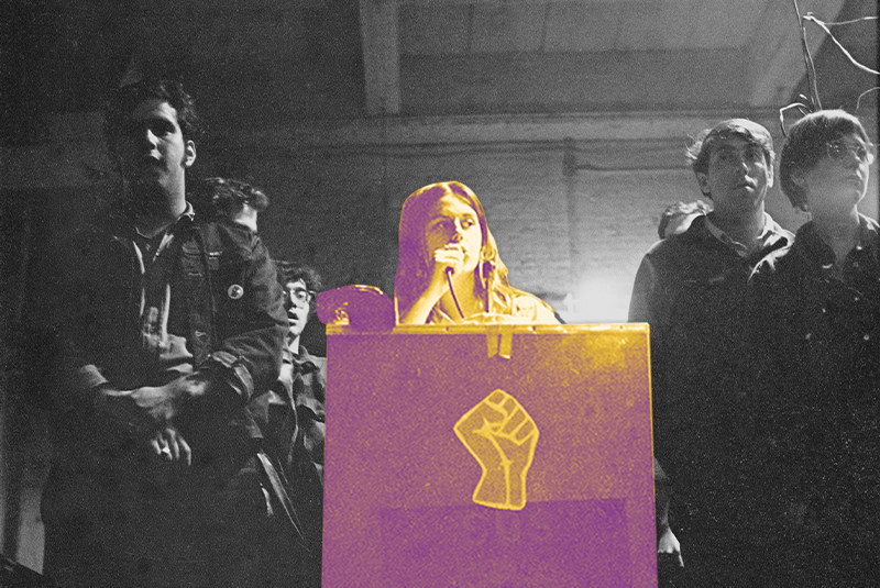 Bernardine Dohrn speaking at a podium with a power fist symbol on the front between six people, all facing a crowd