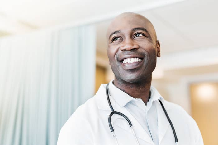 A male doctor smiles while wearing a stethoscope around his neck in a hospital setting