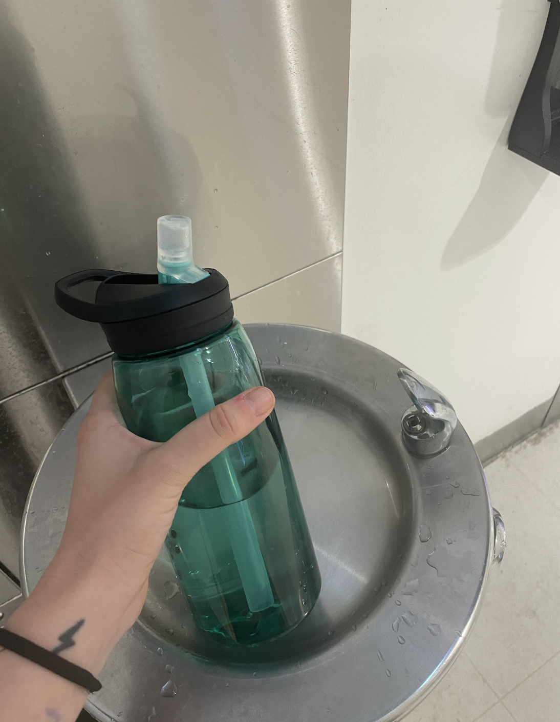 A hand holding a half-full water bottle in front of a drinking fountain