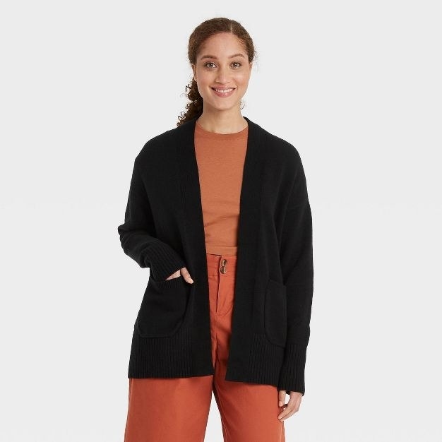 The model wears a black chunky open sweater with an orange shirt and pants