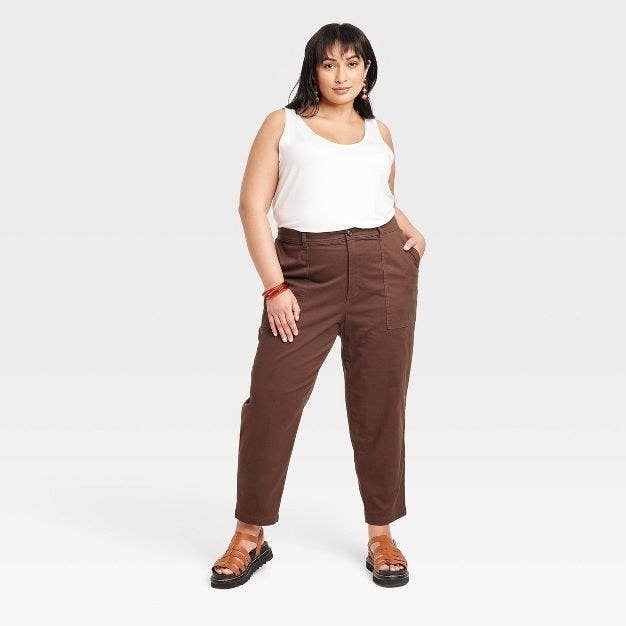 The model wears the dark brown chino pants with a white tank and tan sandals