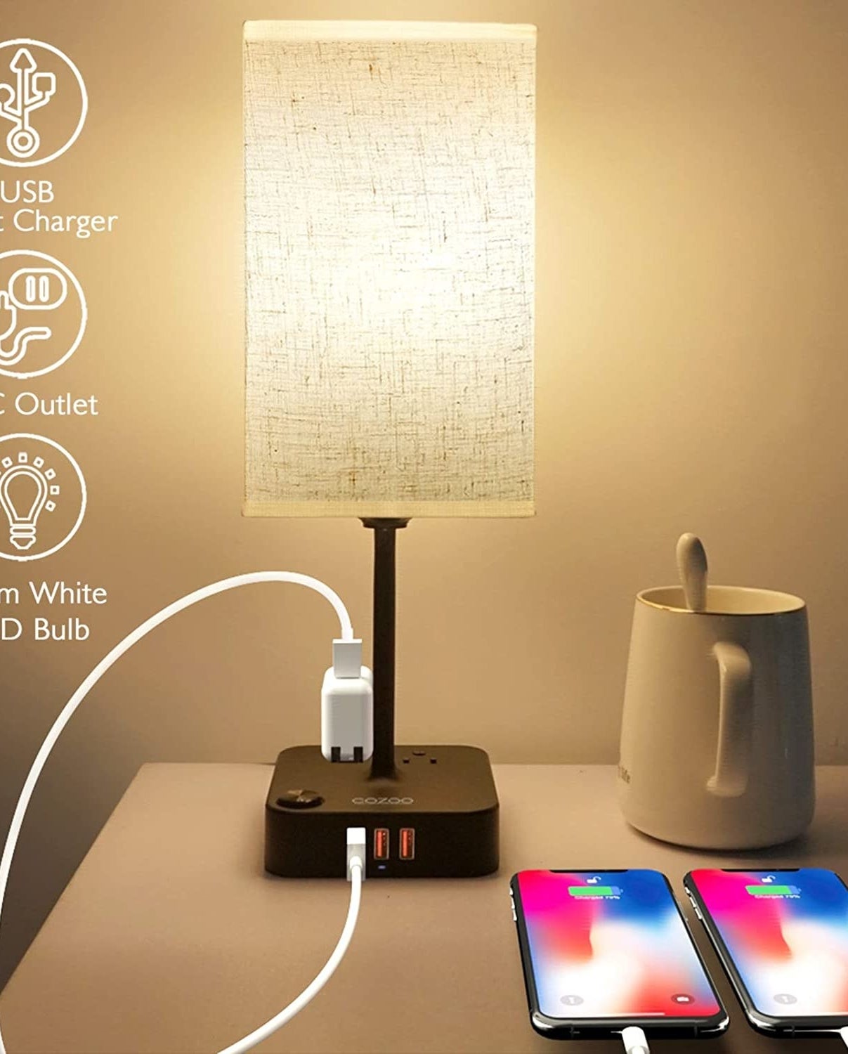 The lamp on a bed side table beside a mug and two charging phones