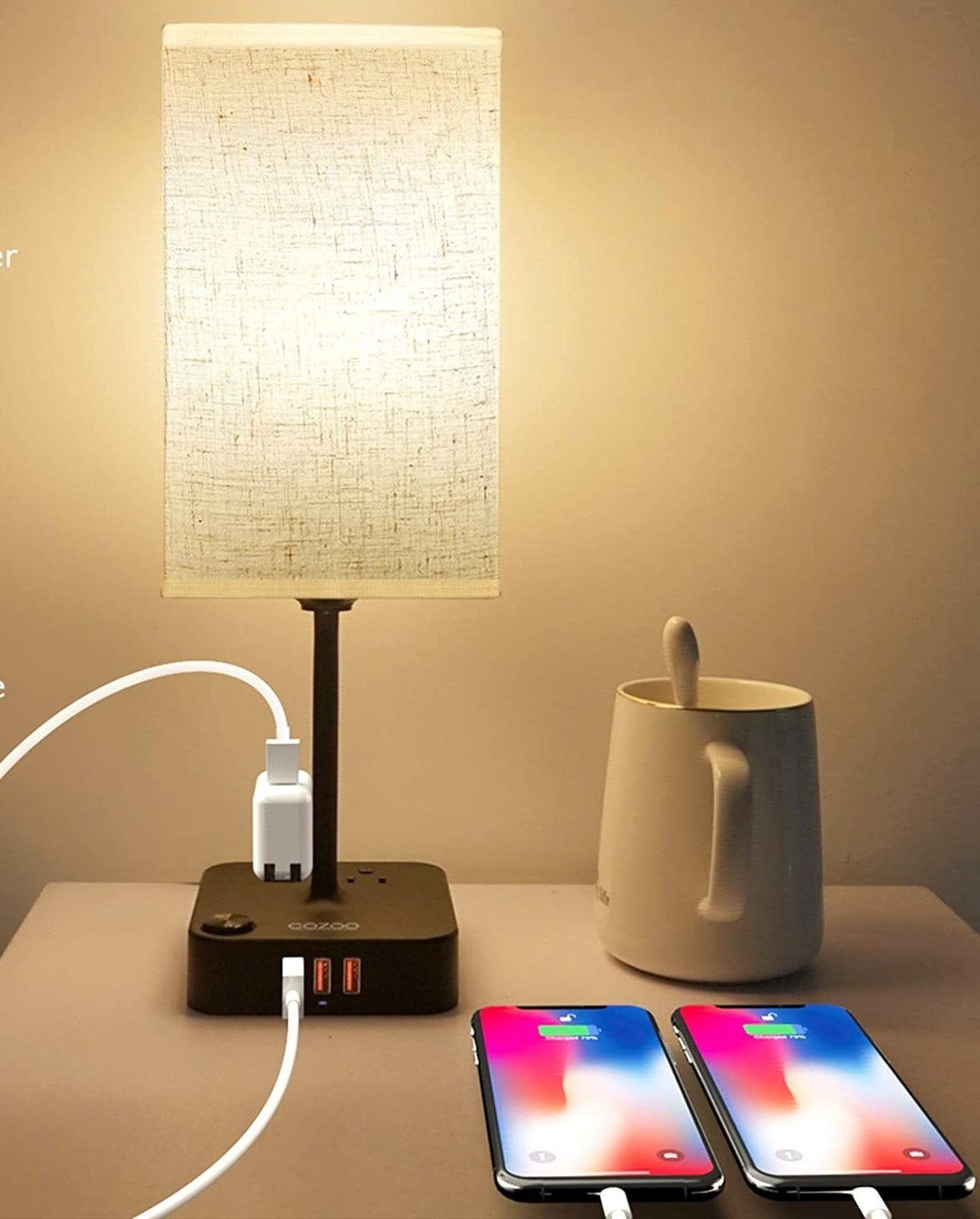 The lamp on a bed side table beside a mug and two charging phones