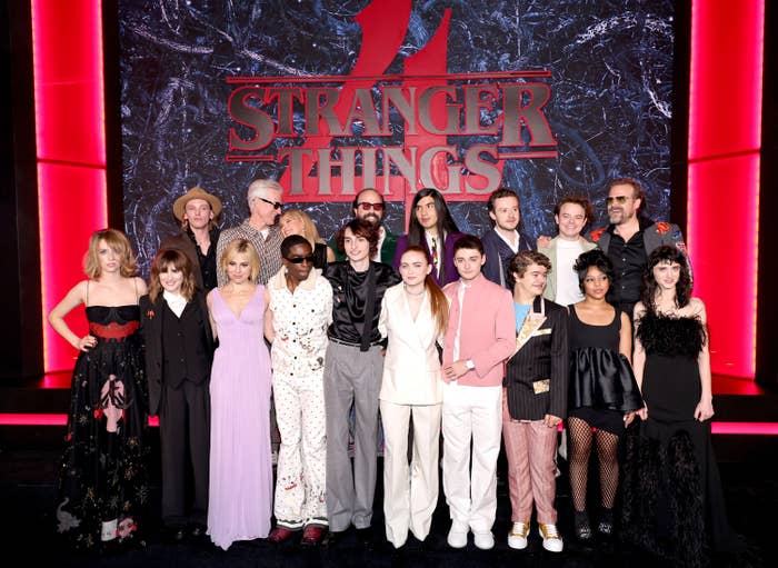 The cast of Stranger Things season 4 smiling at an event, standing in front of backdrop reading &quot;Stranger Things&quot; with a large red number 4 behind it