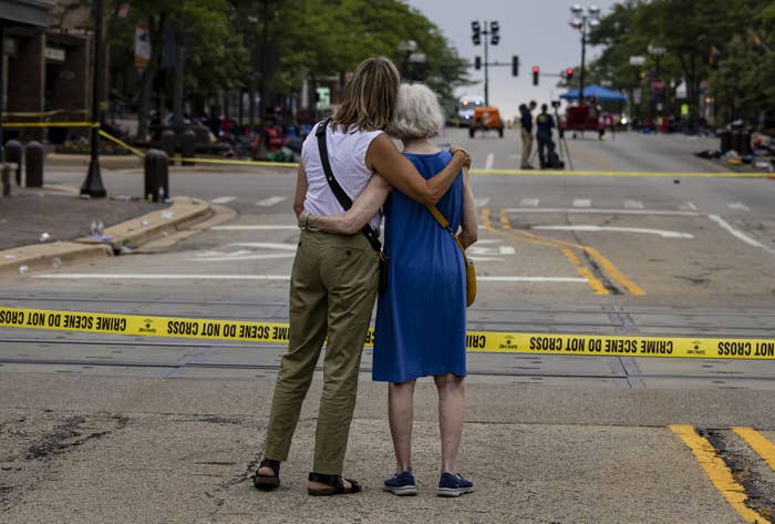 Two women with their backs to the camera hold their arms around each other, facing an empty street where police caution tape is strung up