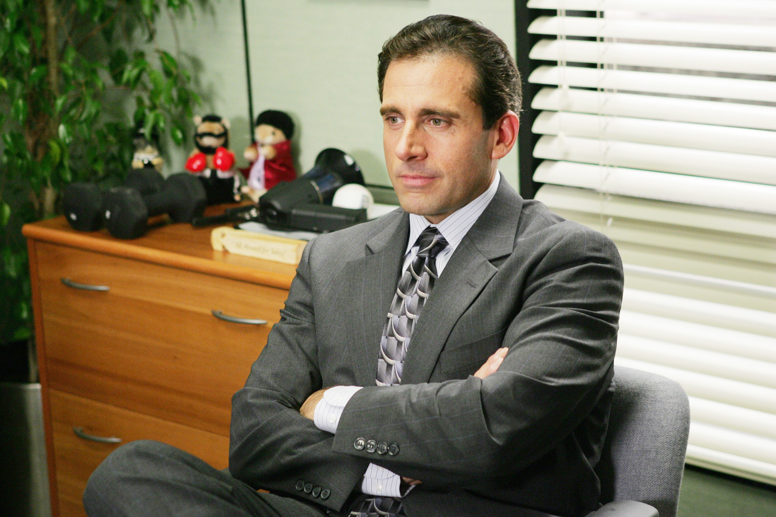 Steve as Michael wearing a suit and sitting with his arms folded
