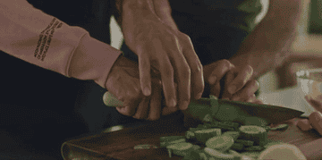 hand holds another hand over a knife guiding it in chopping cucumbers