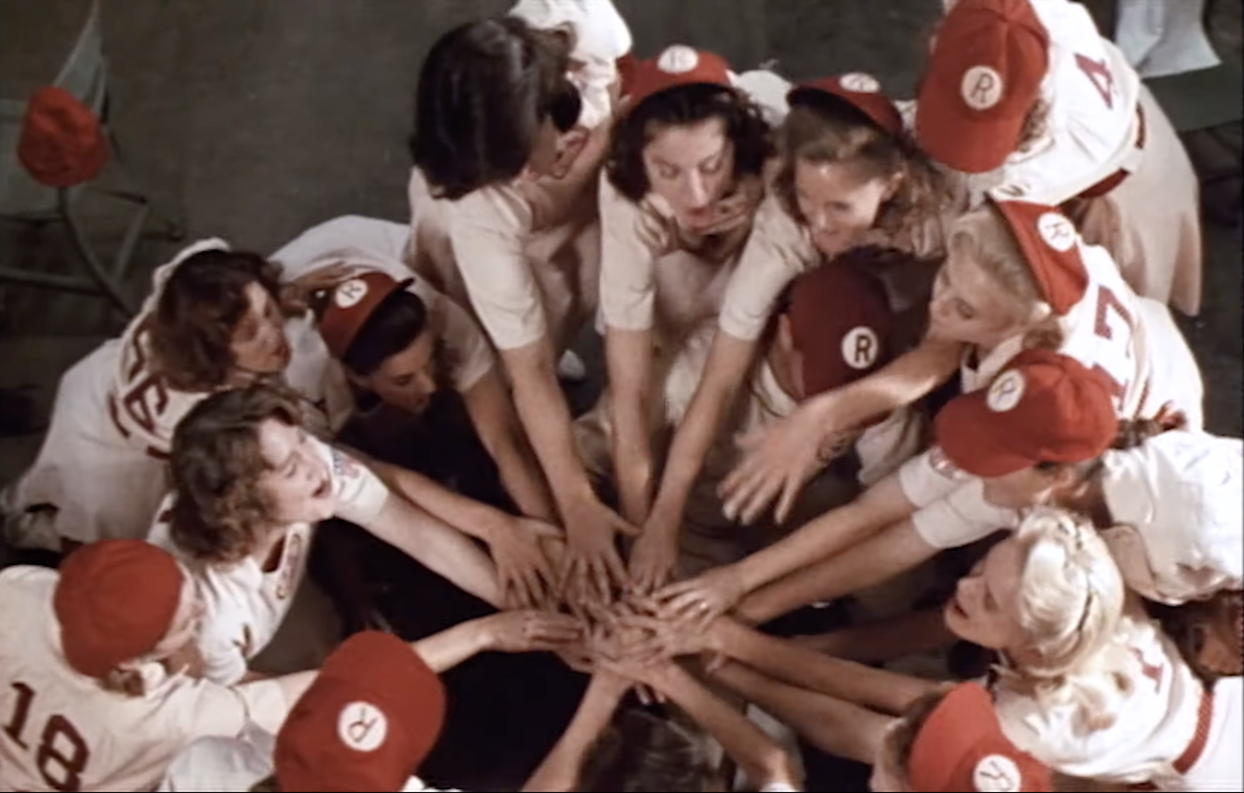 A girls baseball team puts their hands in the center of a huddle.