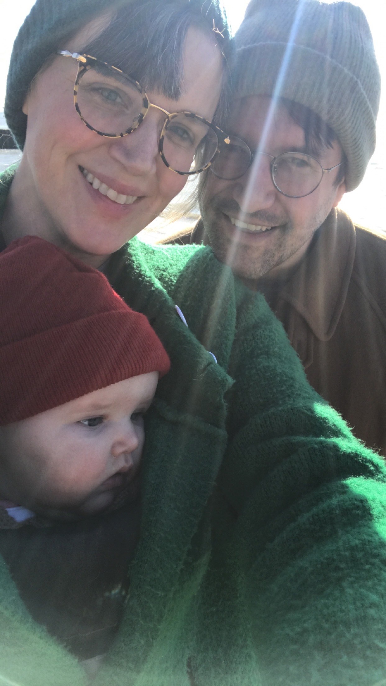 A selfie of Sophie, her husband, Luke, and their daughter outside in cold weather. Sophie is wearing a big green jacket that her daughter is tucked into.