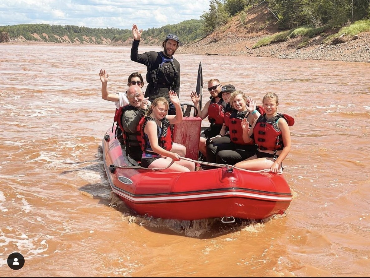 A group of people wearing life vests and waving while aboard a raft on the Shubenacadie River